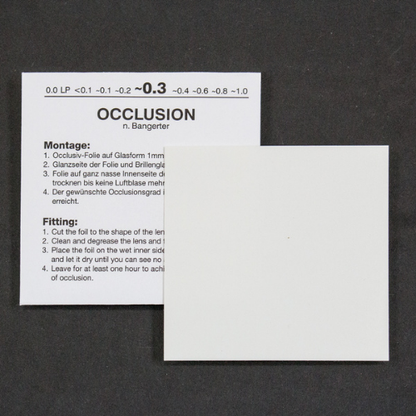 A white instruction card with German nd English text for occlusion lens fitting, shown on a dark textured background with a blank white square overlay in front.