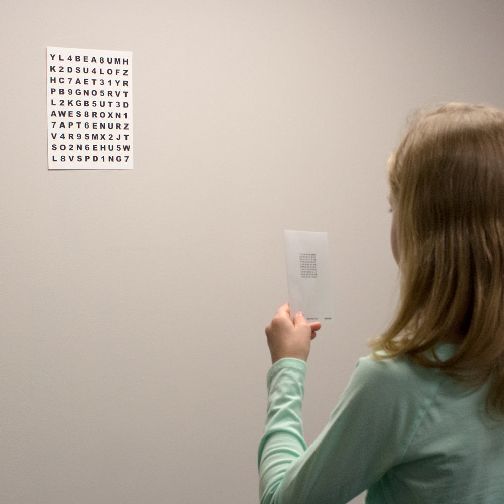 A child in a mint green shirt is shown from behind, holding a small card with text while looking at a white chart of letters and numbers mounted against a wall - testing and accommodative vision training.