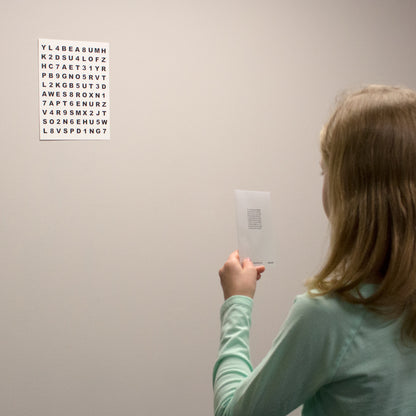 A child in a mint green shirt is shown from behind, holding a small card with text while looking at a white chart of letters and numbers mounted against a wall - testing and accommodative vision training.