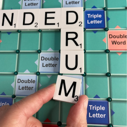 A close-up of a hand placing a Scrabble letter tile on a green grid board, with the word 'DRUM' spelled out in white tiles.