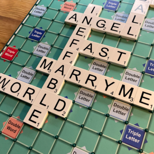 A close-up of a Scrabble game board with letter tiles arranged to spell out ANGEL, FAST, MARRY ME and WORLD across intersecting words. The green grid, double/triple word markers, and wood tabletop are visible around the board.
