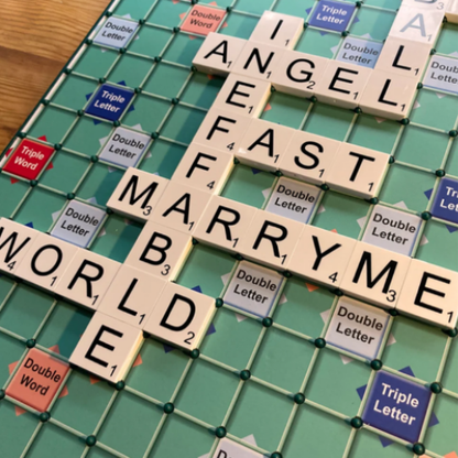 A close-up of a Scrabble game board with letter tiles arranged to spell out ANGEL, FAST, MARRY ME and WORLD across intersecting words. The green grid, double/triple word markers, and wood tabletop are visible around the board.